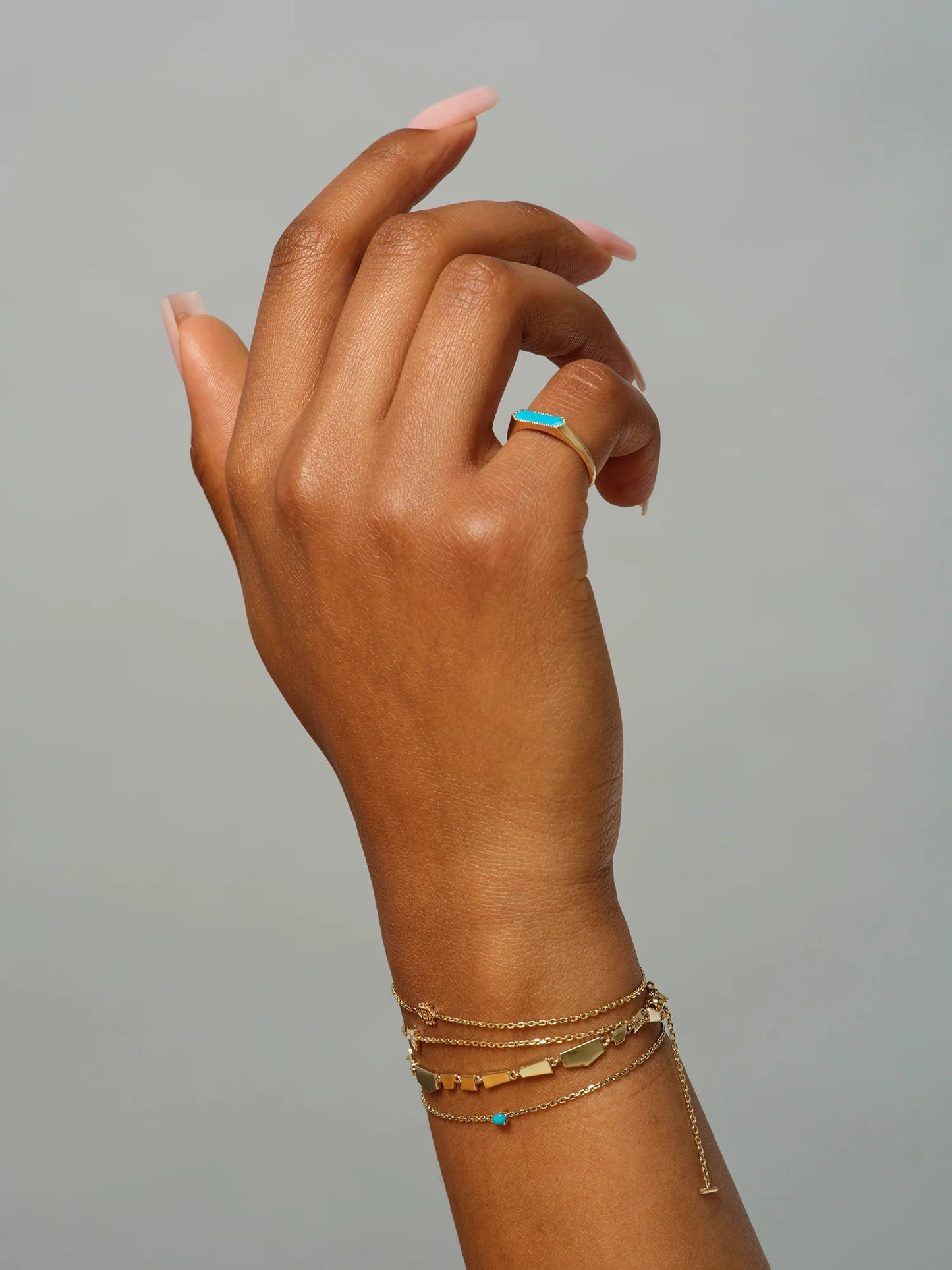 hand model wearing the astra hex turquoise pinky ring on their pinky and four gold chain bracelets on a grey background