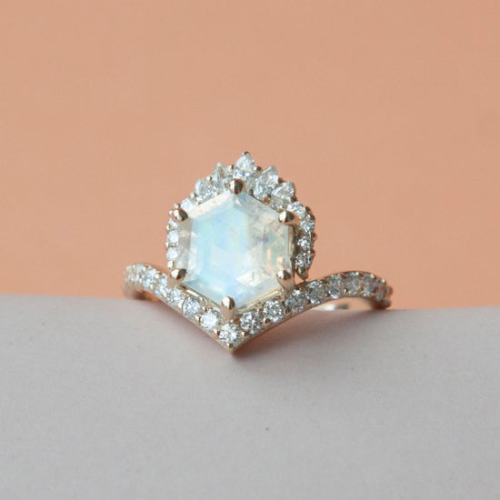 Hexagon moonstone with diamond halo and diamond band ring close up on grey and peach colored background.