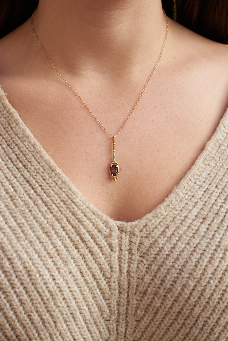 The neck and chest of a light skinned person wearing an oatmeal colored sweater with a V neck and a gold chain with a pendant featuring a rhodolite garnet 