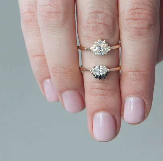 Two sideways set marquise diamond rings, one with a black diamond crown, and one with a white diamond crown, modeled on a hand with grey background.