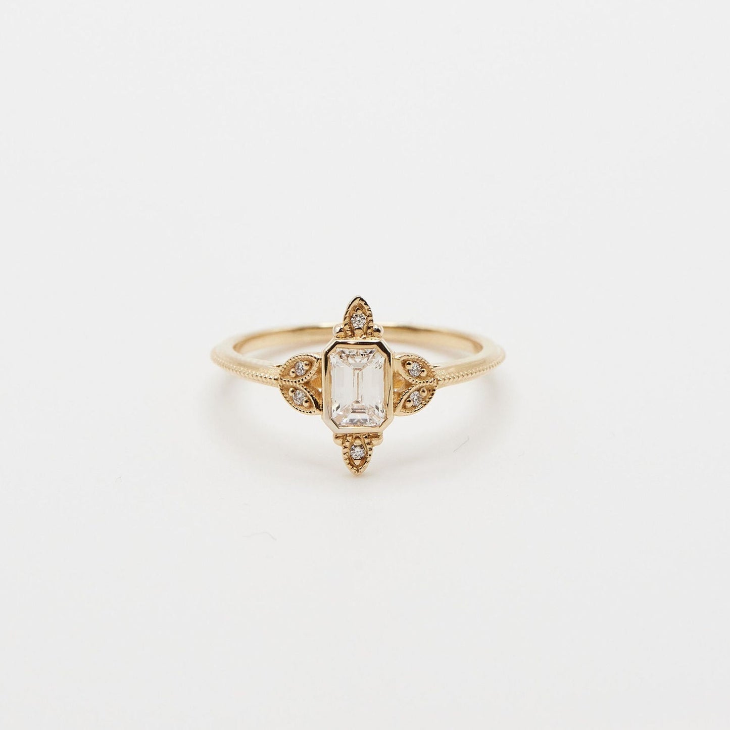 emerald cut diamond ring with milgrain and diamond accents on each side in yellow gold on white background