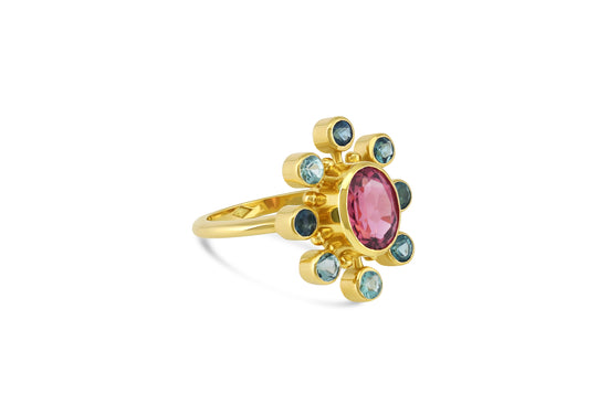 side view of the 18k yellow gold cosmos ring with pink tourmaline center stone and teal tourmaline and gold halo on white background.