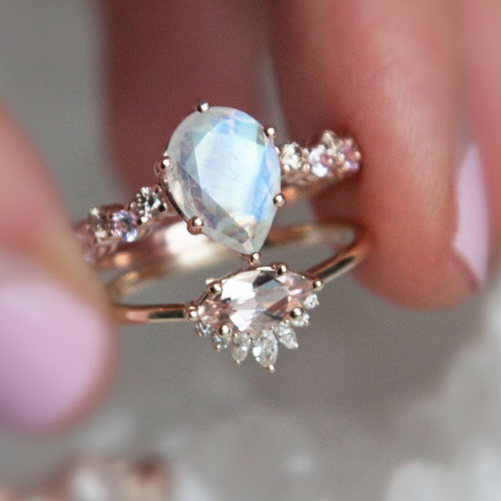 Hand holding pear shaped rainbow moonstone intertwine ring and a morganite and diamond marquise ring, close up.