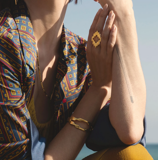 woman in paisley blue, red and yellow blazer pictured in front of the lake wearing a gold geometric ring, as well as a gold bracelet on her left wrist 
