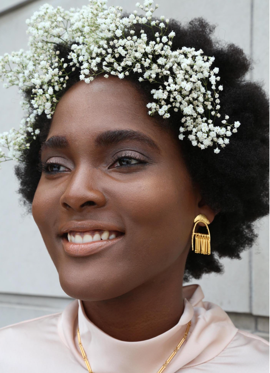 frontal view picture of woman in pink turtle neck wearing white flowers in her hair, gold plated brass dangle earrings worn in her left ear pictured infront of a concrete building