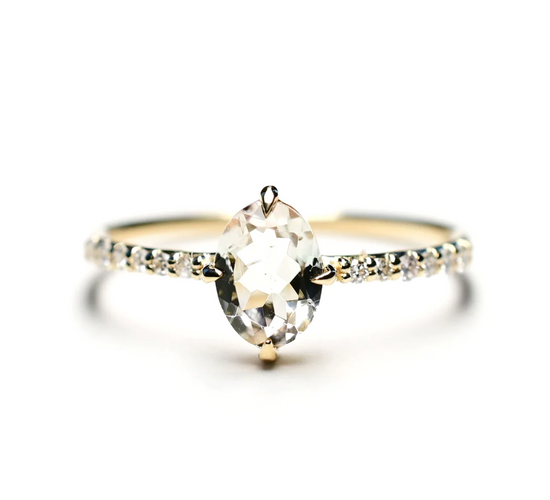 a yellow gold solitaire ring with an oval clear colored tourmaline center stone with white diamonds on the band, sitting on a white background