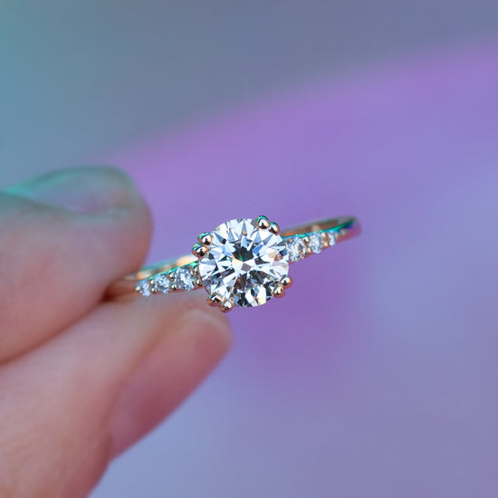 Hand holding diamond solitaire Rheia ring on purple background.