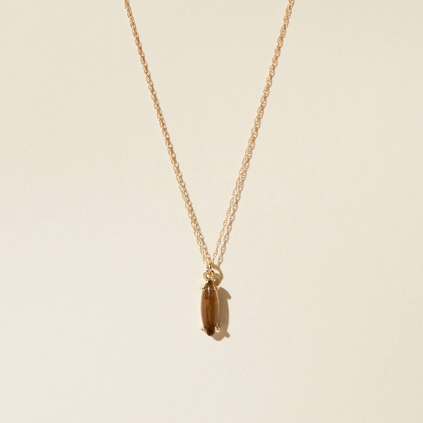 an elongated oval tan glass cabochon prong sent pendant on a gold rope chain on a beige background